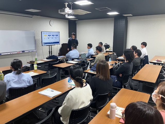 大阪・天神橋筋商店街の会議室で開催したタックス・プラン税理士法人の社内全体会議の様子。プロジェクター前で発表を行う職員と、メモを取りながら参加する社員たち