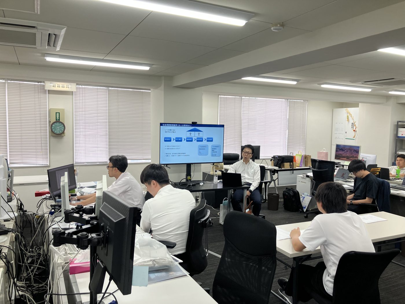 タックス・プラン税理士法人の外部研修の様子。スクリーンに「社会保険 新規適用」の資料を映し、講師が法人設立時の社会保険手続きを解説している場面。