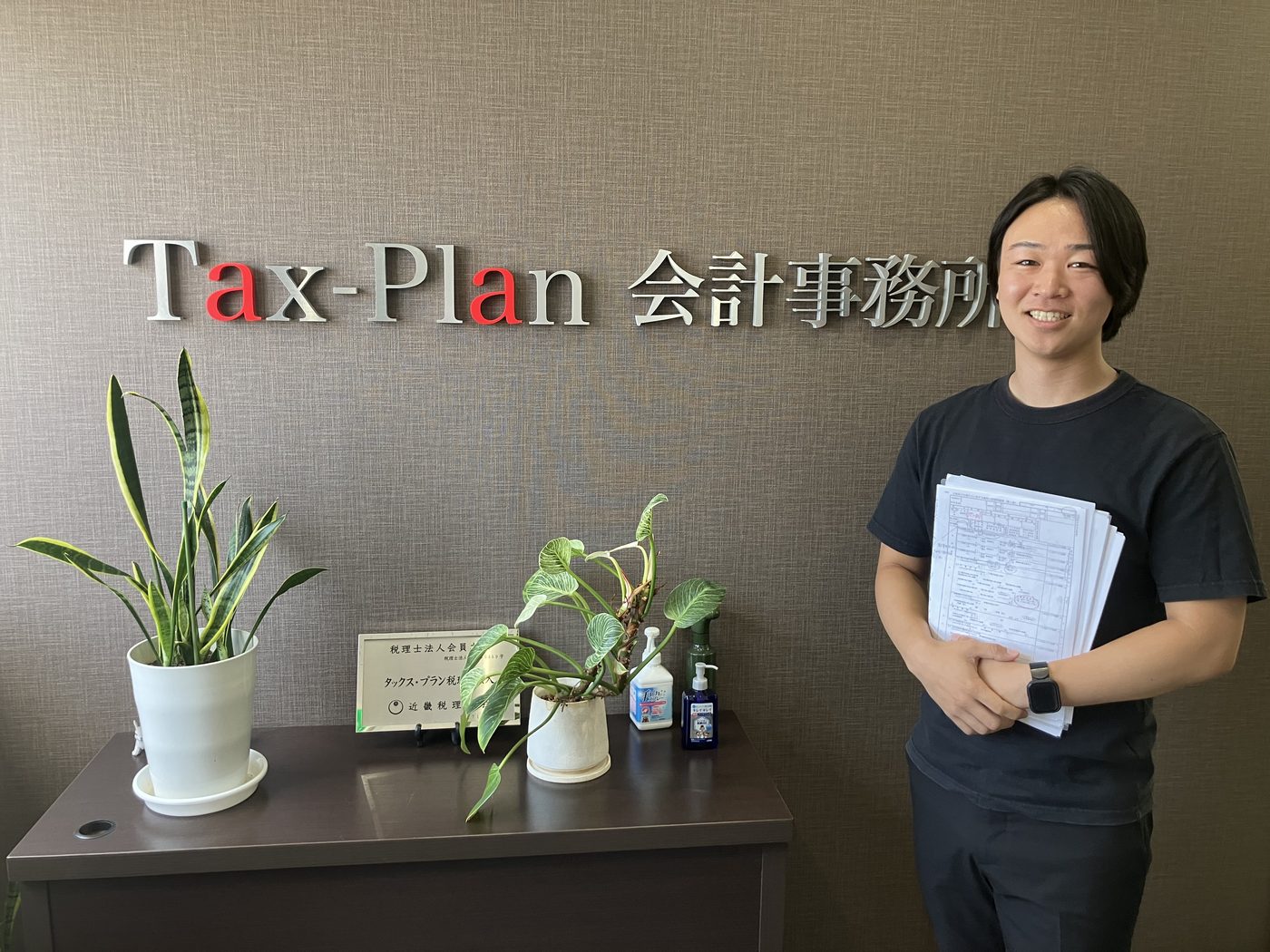 タックス・プラン税理士法人 入社3年目 玉田幹太が担当先の資料を手に事務所エントランスで笑顔で立つ様子